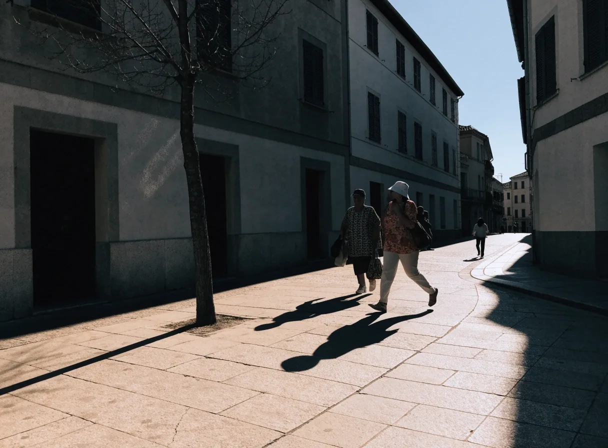 Plaza urbana española a mediodía con dos figuras lejanas y una sombra muy larga sobre el pavimento
